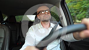 Bearded man in glasses and white shirt driving a car in sunny weather