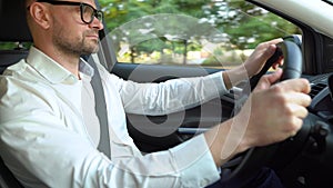 Bearded man in glasses and white shirt driving a car in sunny weather. Side view