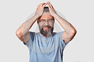 Bearded man in gray t shirt, has round eyewear, keeps hand on head, grimaced, has problems, being in bad mood, has terrible