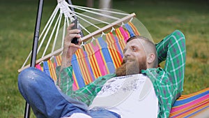 A bearded man in a green checkered shirt lies on a hammock and uses a telephone