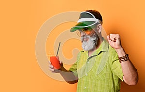 Bearded man in green sun visor, shirt and sunglasses. Holding glass of squeezed juice, clenched fist, posing on orange background