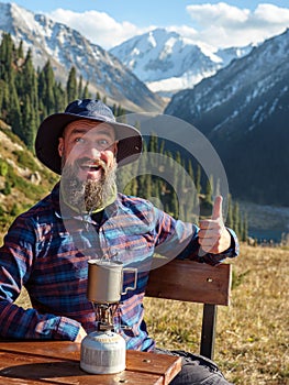 A bearded man heats a drink on a gas burner while outdoors in the mountains