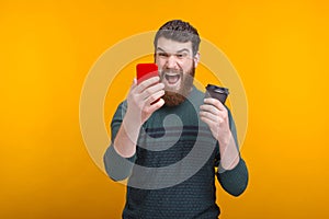 Bearded man holding a black paper cup is shouting at his phone.