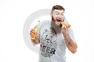 Bearded man holding bottle of beer and eating hot dog