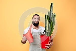 Bearded man holding a watering can and a flower nad looking at the camera
