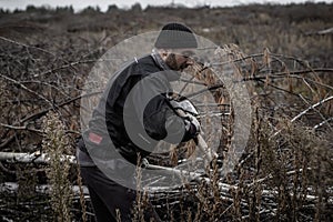 A bearded man holds a trunk of a small tree in his hand, and another raises an ax to strike. A lumberjack cuts off