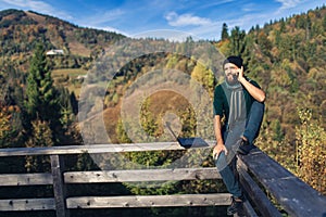 Bearded man with laptop sits on railing and talks on phone. Freelancer hipster on vacation in mountains