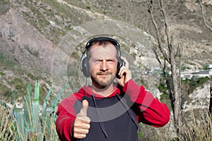 A bearded man listens to music on headphones, in nature. There are mountains behind it. He shows his thumb up.