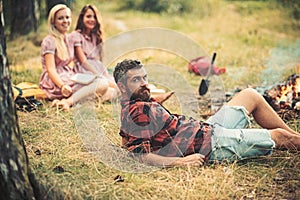 Bearded man in lumberjack shirt lying on grass, unity with nature concept. Group of young hikers camping in forest