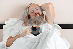 Bearded man lying in cozy bed after waking up holding cup of coffee with his eyes closed enjoying and dreaming