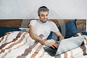 Bearded man lying in morning bed with laptop