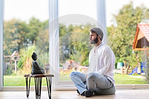 Bearded man meditating cross legged sitting on a pillow in front of a buddha statue.