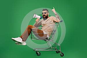 Bearded man with megaphone pointing up while sitting in shopping cart isolated