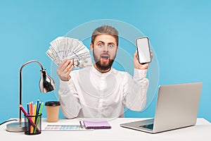 Bearded man office worker looking at camera with astonishment holding big sum of dollar cash and smartphone with white display,