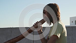 Bearded man playing didgeridoo ustralian instrument on rooftop at sunrise.