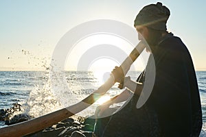 Bearded man playing his didgeridoo Australian instrument in sunshine at seashore.