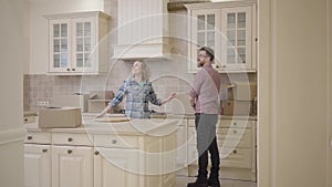 Bearded man and pretty woman enter the kitchen, holding hands. Man carrying box and puts it on table. Woman hugs her