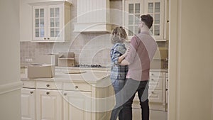 Bearded man and pretty woman enter the kitchen, holding hands. Man puts his wife on the table. Woman hugs her husband