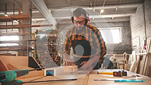 Bearded man putting on headphones and starts cutting a piece of wood with a circular saw