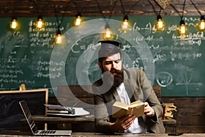 Bearded man read book in classroom. Scientist hipster with book on chalkboard. Businessman in suit read at school desk