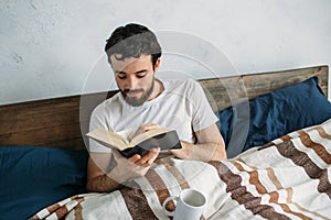 Bearded man reading a big book lying in his bedroom.