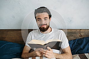 Bearded man reading a big book lying in his bedroom.