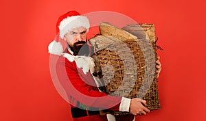 Bearded man in Santa claus costume carrying basket of chopped firewood for fireplace. Winter holidays vacation. Santa