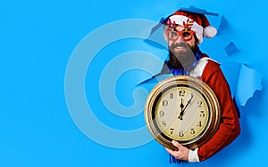 Bearded man in Santa hat and party glasses with clock looking through paper hole. Christmas sales.