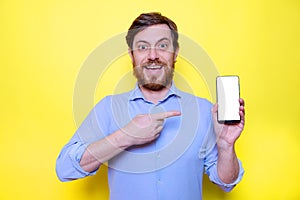 Bearded man showing and pointing at blank screen smartphone on yellow background