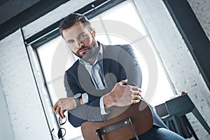 bearded man is sitting on the chair at office and looking very seriously at camera