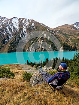 A bearded man sitting on a folding chair rests in the mountains on the shore of a beautiful lake