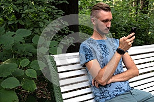 Bearded man sitting on park bench with cellphone, wearing smart watch, background of summer green garden. Hipster male