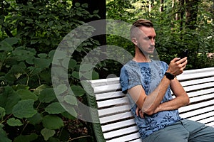 Bearded man sitting on park bench with cellphone, wearing smart watch, background of summer green garden. Hipster male