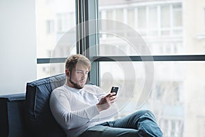 Bearded man sitting on the sofa at the window and using a mobile phone. The office worker resting on the couch