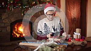 Bearded man sitting and tying a bow on a gifts for New Year near fireplace and looks to camera. Guy wearing Christmas