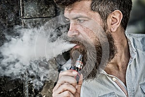 Bearded man smokes vape, white clouds of smoke. Electronic cigarette concept. Man with long beard looks relaxed. Man
