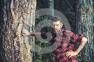 Tall brutal man standing in forest. Lumberjack resting while leaning on tree. Hipster with roses tattoo on arm