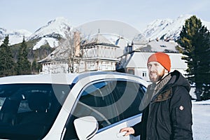 Vousatý muž stojící vedle svého auta na Štrbském Plese s Vysokými Tatrami na pozadí, Slovensko