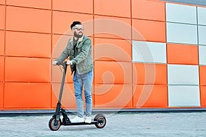 Bearded man in sunglasses posing on electric scooter over orange wall background. Man riding kick scooter.