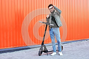 Bearded man in sunglasses posing on electric scooter over orange wall background. Man riding kick scooter.