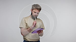 Bearded man in t-shirt thinking, remembering and taking notes in paper notebook, writing creative idea