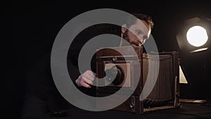 Bearded man takes off lid of old wooden retro accordion camera, adjusts lens under light of lamp in dark isolated room