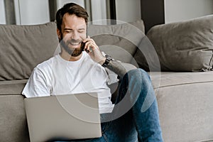 Bearded man talking on cellphone and working with laptop on floor