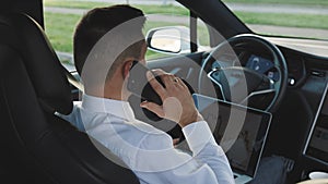 Bearded man talking on smartphone in car with opened laptop on knees. Businessman sitting on seat and fastened with