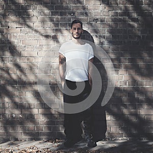 Bearded man with tattoo wearing blank white tshirt and black jeans. Bricks wall background. Square mockup