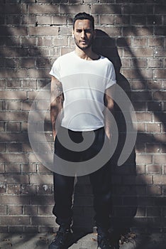 Bearded man with tattoo wearing blank white tshirt and black jeans. Bricks wall background. Vertical mockup, soft