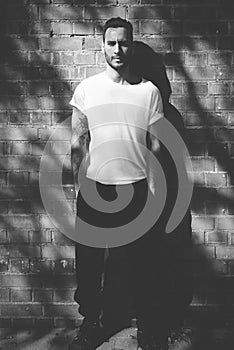 Bearded man with tattoo wearing blank white tshirt and black jeans. Bricks wall background. Vertical mockup, soft