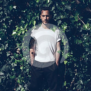 Bearded man with tattoo wearing blank white tshirt and black jeans.Green garden wall background. Square mockup