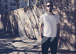 Bearded man with tattoo wearing blank white tshirt and black sunglasses. City garden wall background. Horizontal mockup