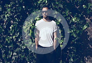 Bearded man with tattoo wearing blank white tshirt and black sunglasses.Green garden wall background. Horizontal mockup